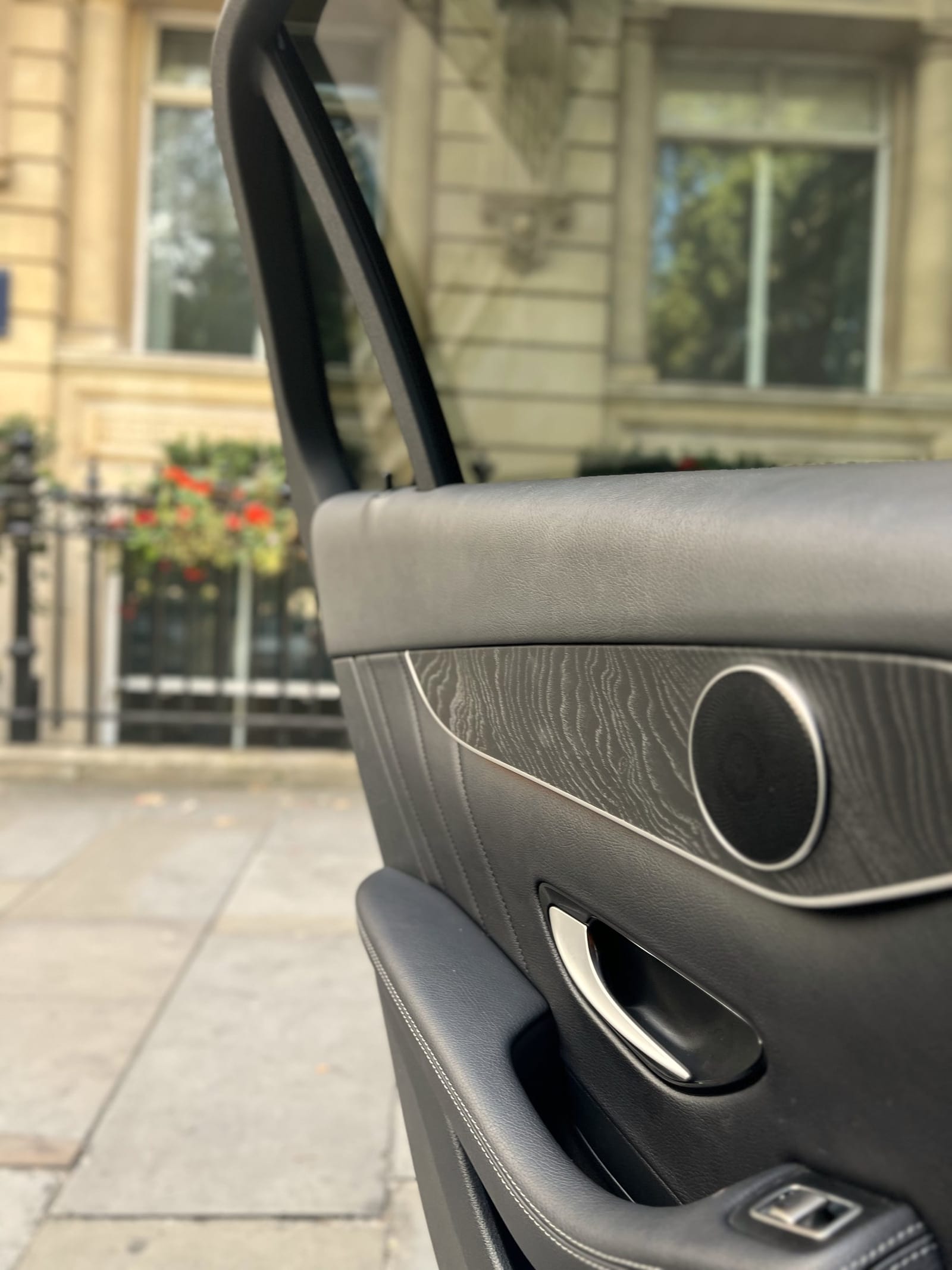 The rear door of an executive car held open beside a stone building with flower-lined railings