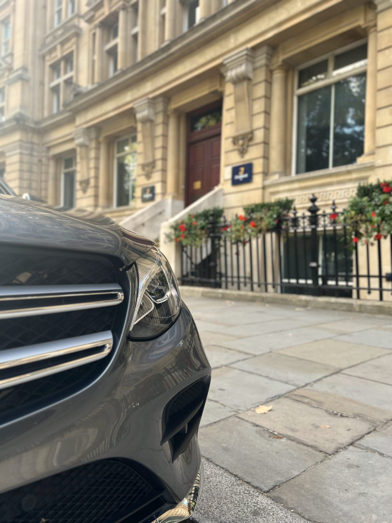 The front of a grey Mercedes parked outside a stone office building with red geraniums