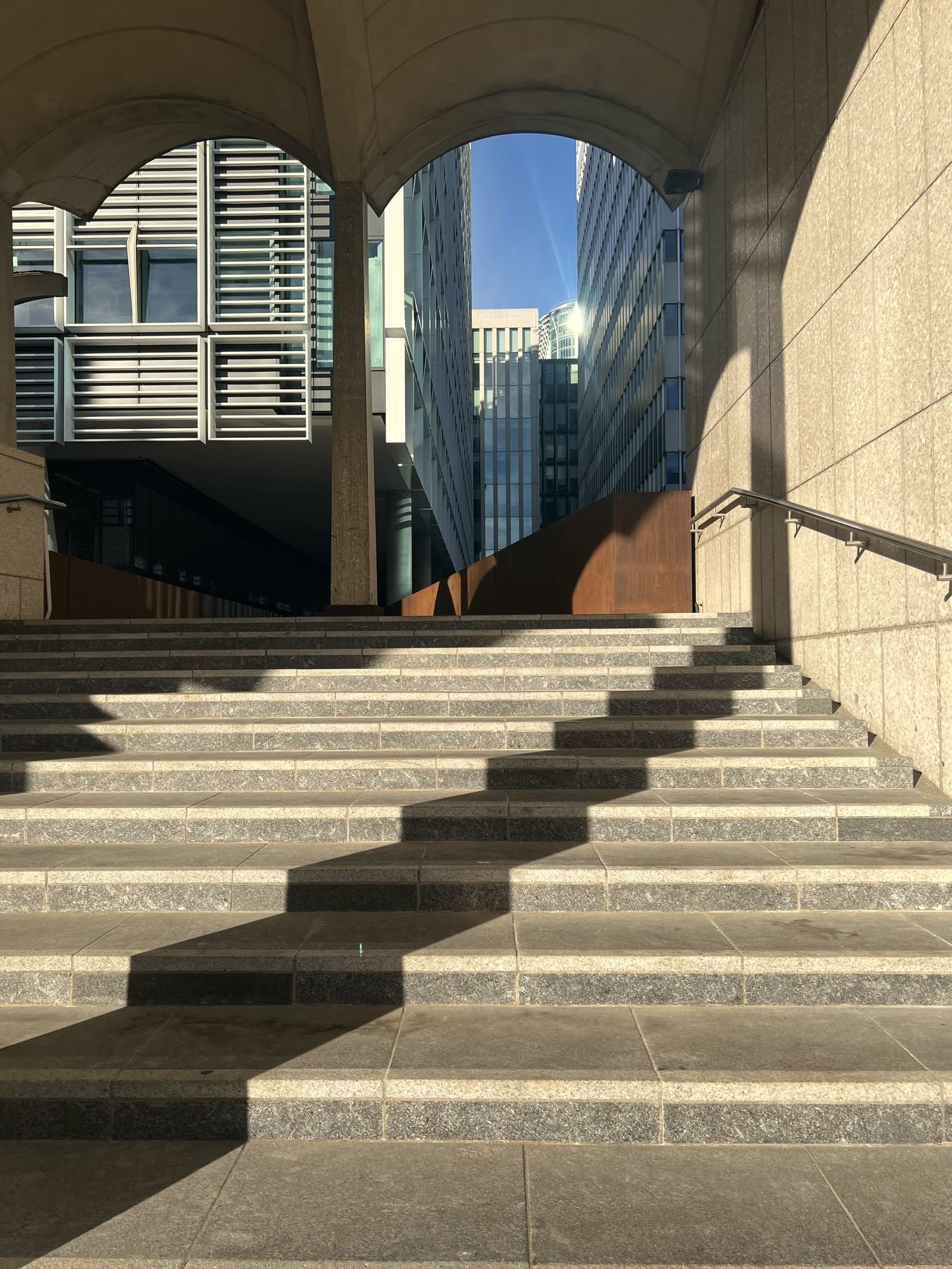 Sunlit steps rising beneath stone arches towards glass office towers
